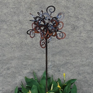 May include: A metal sculpture of a stylized flower with a rusty brown finish. The sculpture is on a tall metal stem and is surrounded by green plants and yellow flowers.