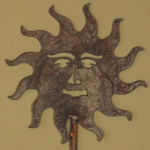 May include: A rustic metal sun face with a smiling expression and radiating rays. The sun has a weathered, textured appearance and is mounted on a thin, rusty metal rod. The sun is a decorative item.