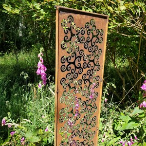 May include: A rusty metal garden screen with a decorative pattern of swirling shapes. The screen is standing in a garden setting with green foliage and pink flowers.