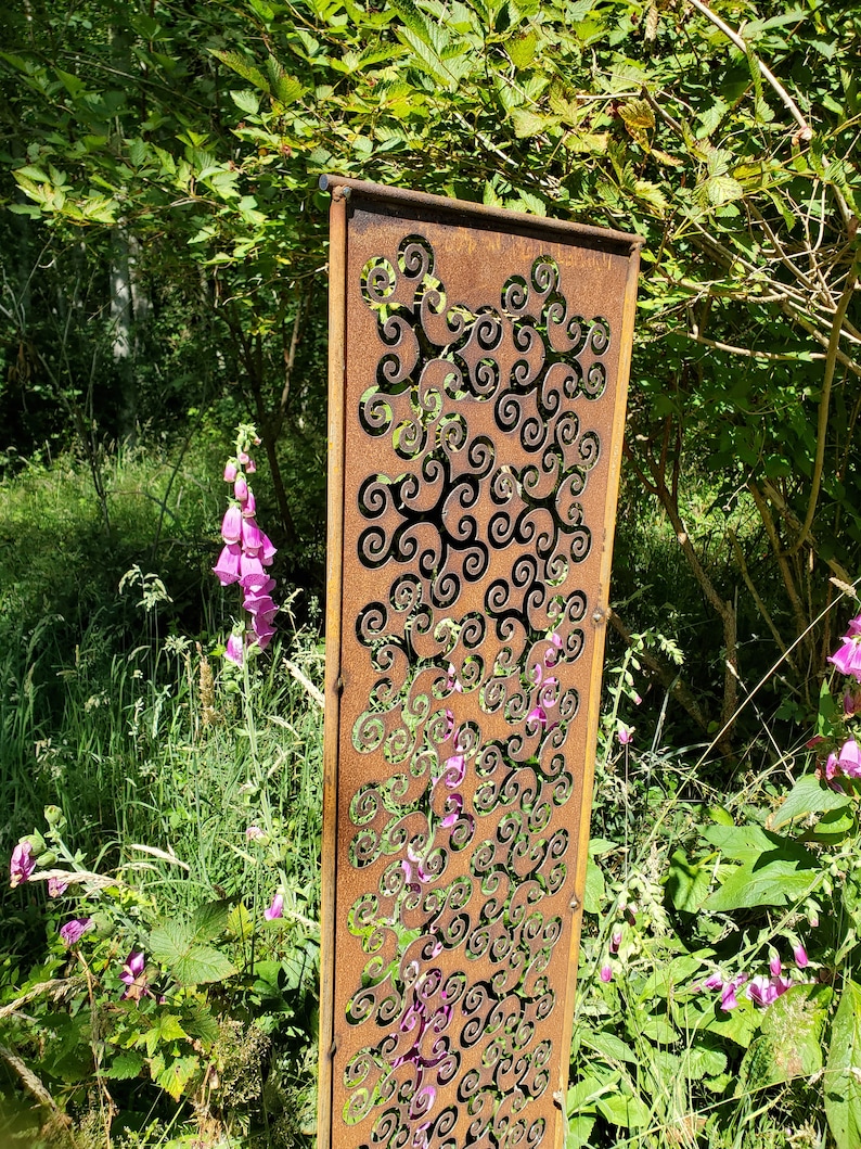 May include: A rusty metal garden screen with a decorative pattern of swirling shapes. The screen is standing in a garden setting with green foliage and pink flowers.