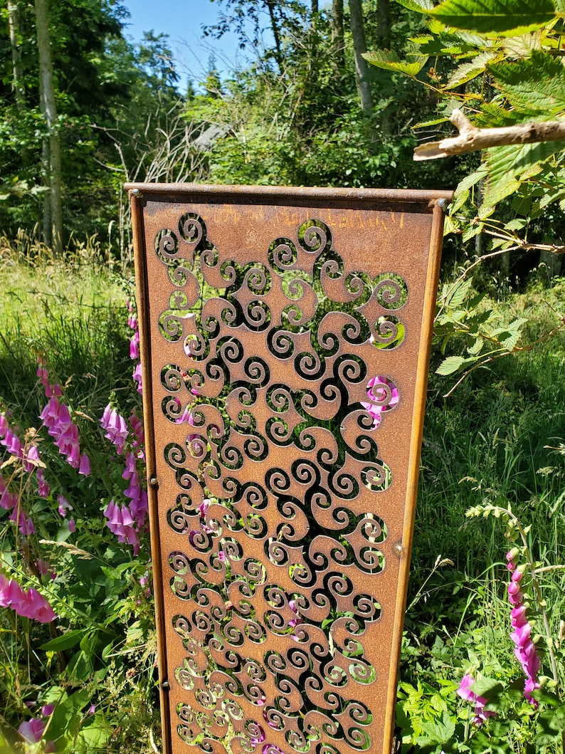 May include: A rusty metal garden screen with a decorative pattern of swirling shapes. The screen is standing in a garden with green grass and pink flowers.
