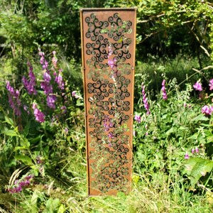 May include: A tall, rusty metal garden screen with a decorative pattern of swirling shapes. The screen is standing in a grassy area with purple flowers.