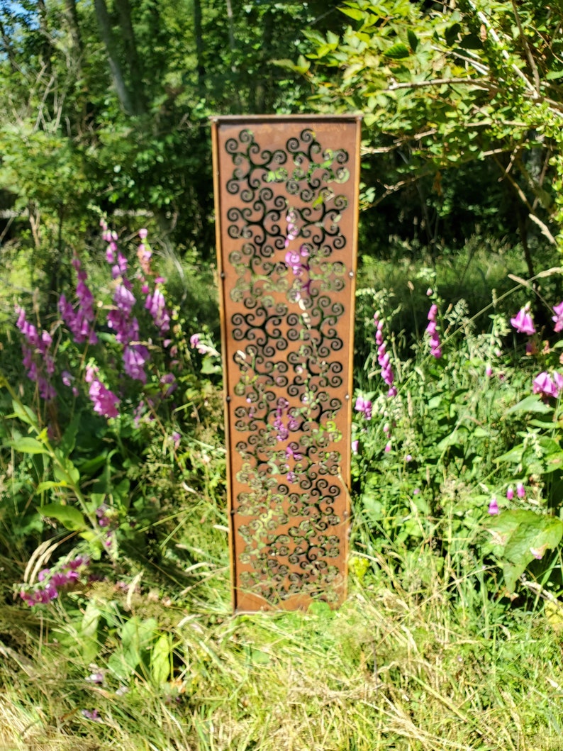 May include: A tall, rusty metal garden screen with a decorative pattern of swirling shapes. The screen is standing in a grassy area with purple flowers.