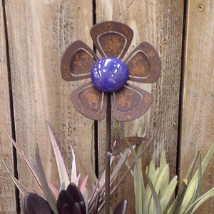 May include: A metal flower garden stake with a rusty brown finish and a blue center. The flower has five petals and is set against a wooden background. The stake is surrounded by artificial plants with purple, pink, and green leaves.