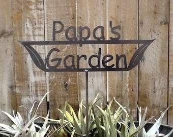May include: A metal garden stake with the words "Papa's Garden" cut out. The stake has a decorative leaf design on each side of the text. The background is a wooden fence, and the stake is placed in a pot with plants.