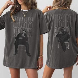 May include: Dark gray t-shirt featuring a black and white image of a person sitting in a chair. The front of the shirt has the text "CHRIS BROWN" and the back has the text "USHER RAYMOND". The shirt is a unisex style.