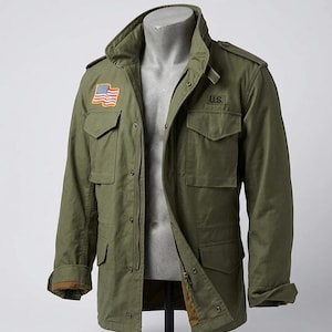 Puede incluir: Chaqueta verde oliva de estilo militar con múltiples bolsillos y cierre de cremallera. Cuenta con un parche de 'U.S.' y un parche de la bandera estadounidense en el pecho. La chaqueta se exhibe en un maniquí.