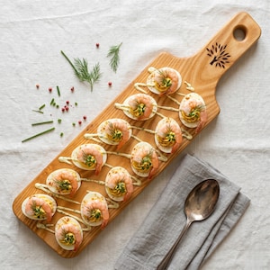 May include: A wooden serving board with a tree design holds devilled eggs topped with prawns and dill. The eggs are arranged in two rows, drizzled with a sauce. A spoon and napkin are on the side. The board is on a white tablecloth.