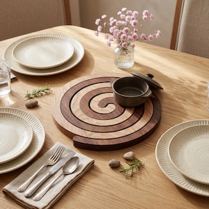 May include: A circular wooden trivet featuring a dark and light wood spiral pattern, placed on a wooden table with cream-colored ribbed plates, a small dark ceramic bowl with a lid, a glass vase with pink flowers, and stainless steel cutlery.