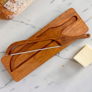 May include: A rectangular wooden cheese slicer board with a matching wooden handle and a thin metal wire cutter. The handle features a small engraved floral detail. The board has a recessed groove for the wire to pass through during use.
