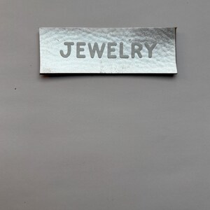 Könnte beinhalten: Ein silbernes Etikett mit dem Wort "JEWELRY" in weiß geprägt.