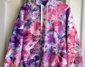 May include: A tie-dye hoodie in shades of pink, purple, and white. The sweatshirt has a front pocket, a hood with a drawstring, and long sleeves. The garment is hanging on a wooden hanger.