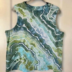 May include: A sleeveless top with a tie-dye pattern in shades of green, blue, and white. The design features wavy lines and a round neckline. The shirt is hanging on a black hanger.