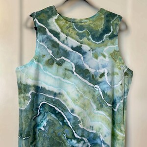 May include: A sleeveless top with a tie-dye pattern in shades of green, blue, and white. The fabric has a marbled effect, with flowing lines of color. The top is hanging on a black hanger against a neutral background.