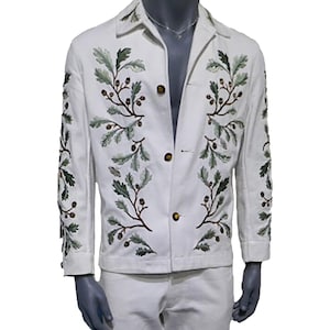 Puede incluir: Una chaqueta blanca con botones y mangas largas. La chaqueta está bordada con hojas verdes y bellotas marrones.