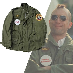 Peut inclure: Une veste de style militaire verte avec une fermeture éclair et plusieurs poches. La veste a trois patchs cousus sur le devant : un avec une tête de mort et des os croisés, un avec les mots "We Are The People" et un avec un avion.