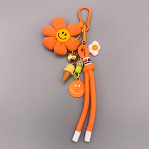 Könnte beinhalten: Ein orangefarbener Schlüsselanhänger mit einer Plüschblume mit gelbem Smiley-Gesicht, einem goldenen Herzanhänger, einer Miniatur-Eistüte, einem kleinen Spiegelei-Anhänger und einem runden orangefarbenen Smiley-Tag an einer langen orangefarbenen Kordel.