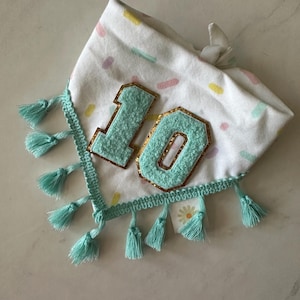 May include: White fabric bandana with a pastel sprinkle pattern and the number "10" in light blue with gold trim. The bandana is trimmed with a turquoise crochet edge and turquoise tassels.