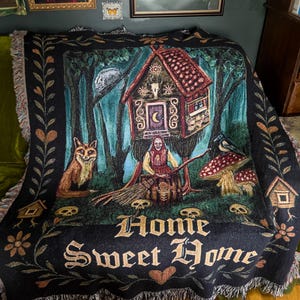 May include: A black woven throw blanket featuring a central illustration of a house on chicken legs, a figure with a broom, a fox, a crow, mushrooms, and skulls. The text "Home Sweet Home" is printed in a gothic font at the bottom with fringe edges.
