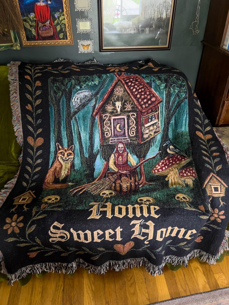 May include: A black woven throw blanket featuring a central illustration of a house on chicken legs, a figure with a broom, a fox, a crow, mushrooms, and skulls. The text "Home Sweet Home" is printed in a gothic font at the bottom with fringe edges.