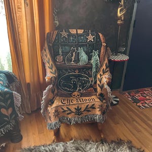 May include: An upholstered armchair with a decorative throw featuring a celestial and feline design. The throw displays a window scene with cats, stars, and the text "The Moon." The chair has wooden arms and a fringed edge.