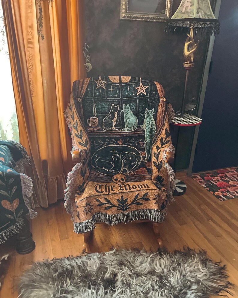 May include: An upholstered armchair with a decorative throw featuring a celestial and feline design. The throw displays a window scene with cats, stars, and the text "The Moon." The chair has wooden arms and a fringed edge.