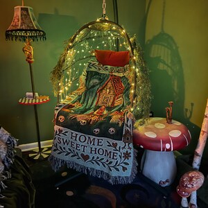 May include: A hanging egg chair draped with a woven throw blanket featuring a gingerbread house, skulls, and the text HOME SWEET HOME. The chair is illuminated by string lights and accented with greenery, positioned near a mushroom-shaped side table.