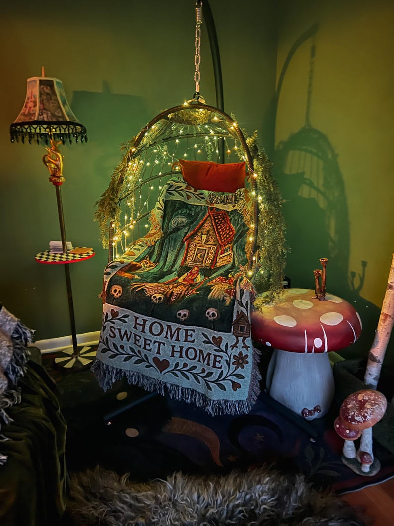 May include: A hanging egg chair draped with a woven throw blanket featuring a gingerbread house, skulls, and the text HOME SWEET HOME. The chair is illuminated by string lights and accented with greenery, positioned near a mushroom-shaped side table.
