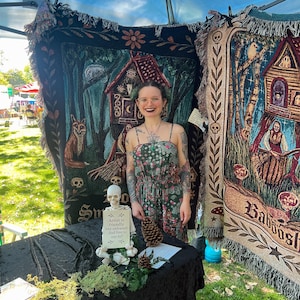 May include: Two large woven tapestry blankets featuring dark forest and witchy illustrations hang behind a table. A small plastic skeleton sits on the black table holding a sign that reads: Artist is friendly but awkward-feel free to say hi!