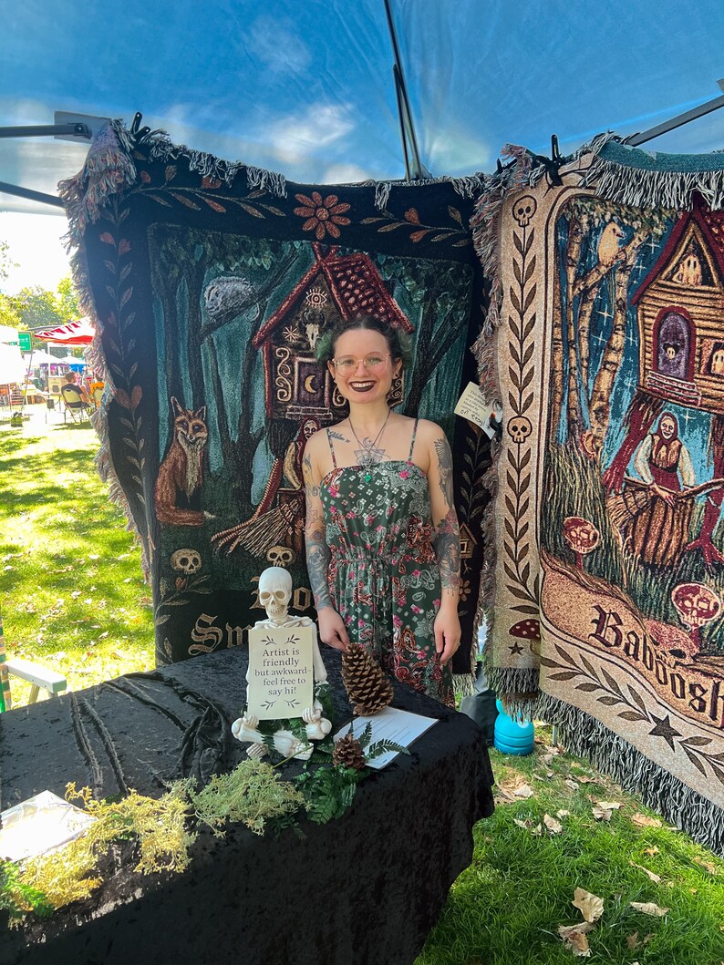 May include: Two large woven tapestry blankets featuring dark forest and witchy illustrations hang behind a table. A small plastic skeleton sits on the black table holding a sign that reads: Artist is friendly but awkward-feel free to say hi!