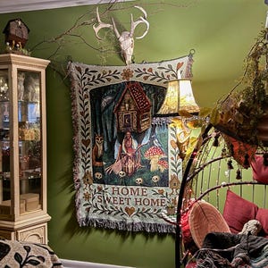 May include: A woven wall tapestry featuring a central illustration of a figure with a broomstick, a house on stilts, mushrooms, and a fox, with the text HOME SWEET HOME at the bottom, framed by a floral border with hearts and small house icons.