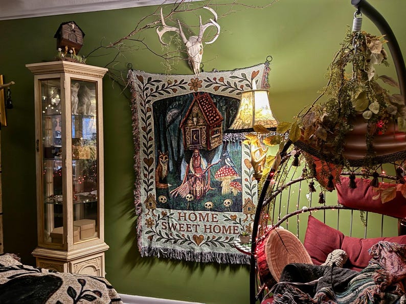 May include: A woven wall tapestry featuring a central illustration of a figure with a broomstick, a house on stilts, mushrooms, and a fox, with the text HOME SWEET HOME at the bottom, framed by a floral border with hearts and small house icons.