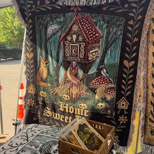 May include: A black woven throw blanket featuring a cottage on chicken legs, a figure with a broom, a fox, a bird, mushrooms, and skulls. The text "Homie Sweet" is woven at the bottom. The blanket has a fringed edge and a decorative floral border.