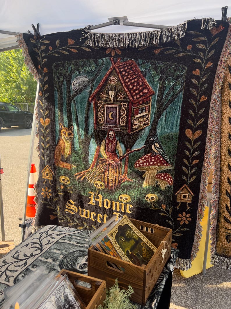 May include: A black woven throw blanket featuring a cottage on chicken legs, a figure with a broom, a fox, a bird, mushrooms, and skulls. The text "Homie Sweet" is woven at the bottom. The blanket has a fringed edge and a decorative floral border.