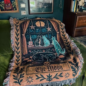 May include: A woven throw blanket featuring a celestial design with cats, a dog, and the text "The Moon." The blanket has a tan background with teal, brown, and black imagery. The design includes a window scene with stars and a crescent moon.