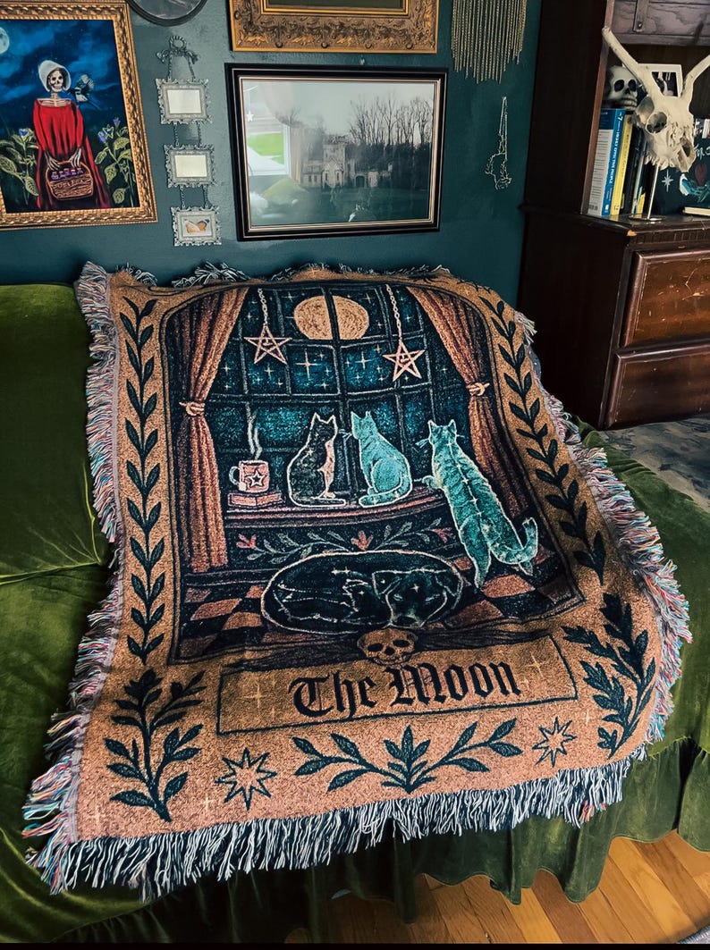 May include: A woven throw blanket featuring a celestial design with cats, a dog, and the text "The Moon." The blanket has a tan background with teal, brown, and black imagery. The design includes a window scene with stars and a crescent moon.
