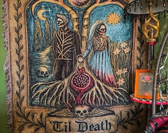 May include: A woven throw blanket featuring a gothic illustration of two skeletal figures holding hands. The design includes a pomegranate, a skull, a crescent moon, a sun, and the text 'Til Death' centered at the bottom in a dark font.