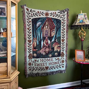 May include: A rectangular woven tapestry featuring a central illustration of a figure with a broom, a fox, a bird, mushrooms, and a house in a forest. The border includes floral motifs and skulls. The text HOME SWEET HOME is printed at the bottom.
