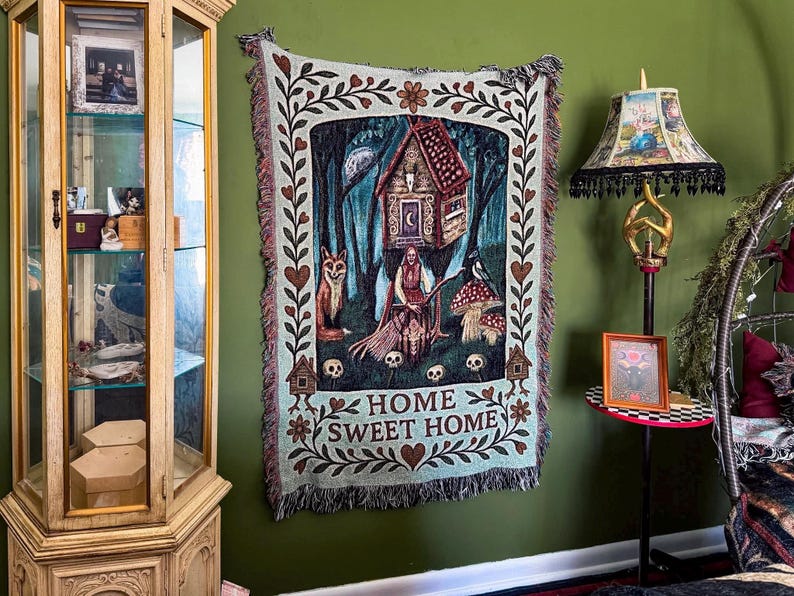 May include: A rectangular woven tapestry featuring a central illustration of a figure with a broom, a fox, a bird, mushrooms, and a house in a forest. The border includes floral motifs and skulls. The text HOME SWEET HOME is printed at the bottom.