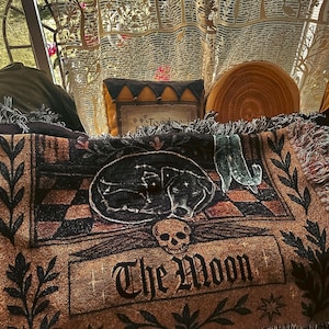 May include: A brown and black throw blanket with a sleeping dog design, the words "The Moon", and a skull. The blanket has a fringed edge and is decorated with stars and floral elements. A decorative pillow and a wooden slice are also visible.