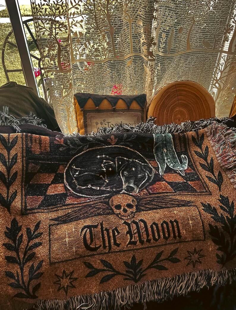 May include: A brown and black throw blanket with a sleeping dog design, the words "The Moon", and a skull. The blanket has a fringed edge and is decorated with stars and floral elements. A decorative pillow and a wooden slice are also visible.