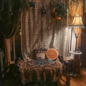 May include: A cozy, dimly lit room with a mystical theme. Features include a patterned curtain, a decorative throw blanket with "The Moon" text, a crescent moon decoration, and a floor lamp with a patterned lampshade. The room is decorated with stars and plants.