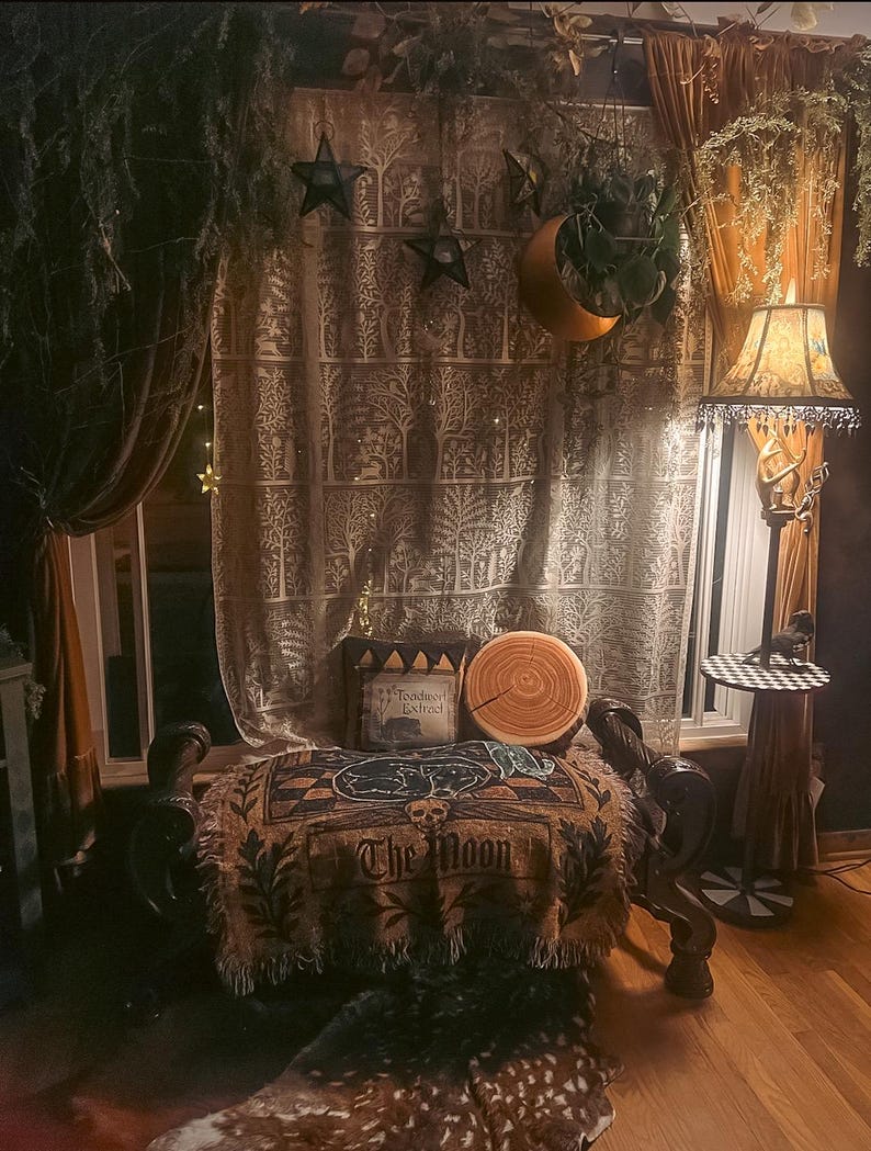May include: A cozy, dimly lit room with a mystical theme. Features include a patterned curtain, a decorative throw blanket with "The Moon" text, a crescent moon decoration, and a floor lamp with a patterned lampshade. The room is decorated with stars and plants.