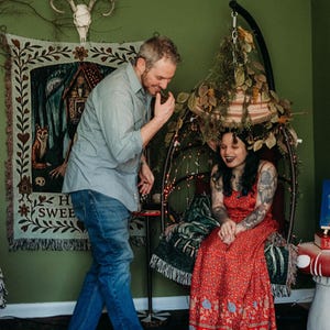 May include: A light blue button-down shirt and blue jeans worn by one individual, and a red floral print maxi dress worn by another individual sitting in a hanging chair decorated with dried foliage and string lights, near a wall tapestry.