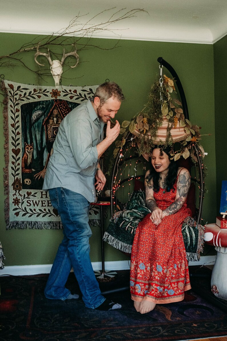 May include: A light blue button-down shirt and blue jeans worn by one individual, and a red floral print maxi dress worn by another individual sitting in a hanging chair decorated with dried foliage and string lights, near a wall tapestry.