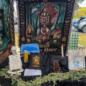 May include: A woven tapestry featuring a witch, a house on chicken legs, a fox, and a raven with the text Home Sweet Home. A skeleton figurine holds a sign reading Artist is friendly but awkward- feel free to say hi! next to a framed print.