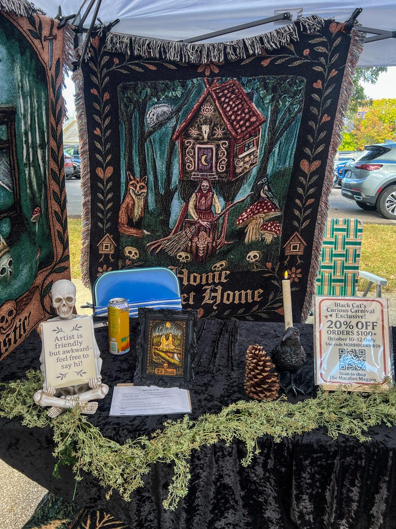May include: A woven tapestry featuring a witch, a house on chicken legs, a fox, and a raven with the text Home Sweet Home. A skeleton figurine holds a sign reading Artist is friendly but awkward- feel free to say hi! next to a framed print.