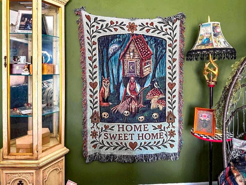 May include: A rectangular woven tapestry featuring a central illustration of a house on stilts, a figure with a broom, a fox, a bird, and mushrooms, surrounded by a floral border with hearts. The text HOME SWEET HOME is printed at the bottom.