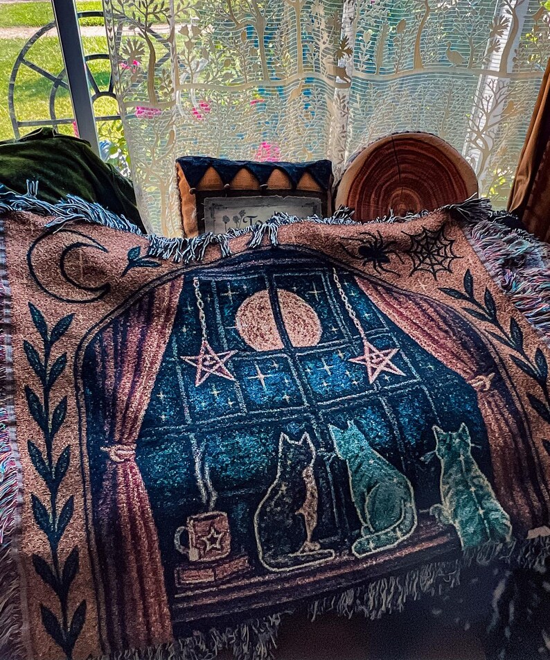 May include: A woven throw blanket featuring a nighttime scene with three cats looking out a window at the moon and stars. The blanket has a fringe and a color palette of blue, brown, and pink.