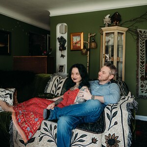 May include: A person in a red patterned maxi dress and a person in a blue button-down shirt and blue jeans sitting on a couch covered with a cream-colored throw blanket featuring a black floral print and a pillow with the text Crows Gather Here.